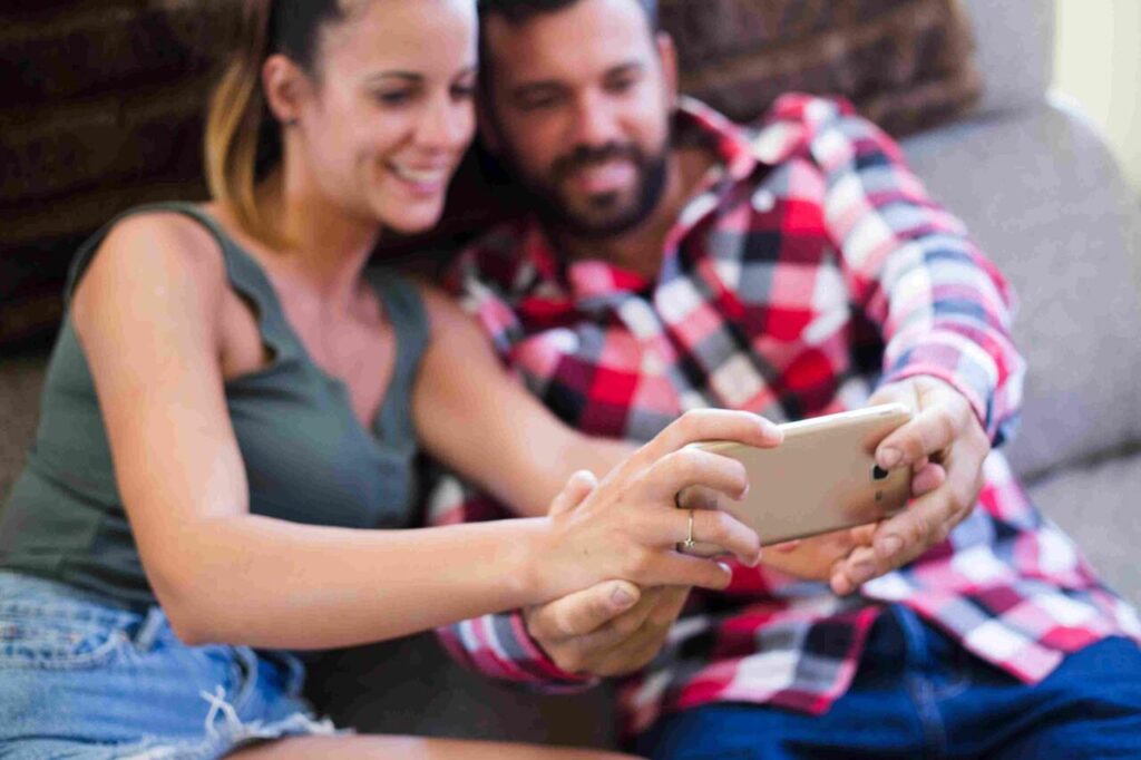 Um casal está sentado de forma relaxada em um sofá cinza claro. A mulher, à esquerda, veste uma regata verde-oliva e shorts jeans. O homem, à direita, usa uma camisa xadrez vermelha e preta e calça jeans. Ambos estão sorrindo e olhando juntos para a tela de um smartphone dourado, que o homem segura com as duas mãos e a mulher apoia com uma mão. Eles parecem estar conferindo o resultado de sua aposta múltipla. A imagem tem um foco suave.