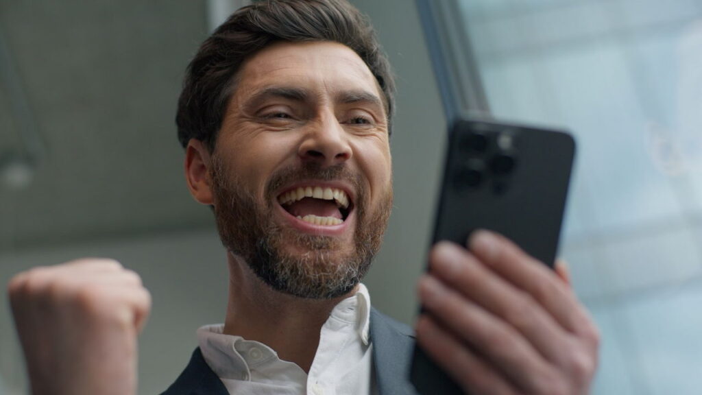Homem jovem com barba, vestindo camisa social e terno, olha para a tela de um smartphone com expressão de extrema alegria e empolgação. Ele está com a boca aberta em um grito de vitória e o punho cerrado, celebrando um ganho ou bônus ao aplicar dicas no Big Bass Bonanza slot. O plano de fundo mostra uma estrutura moderna de vidro.
