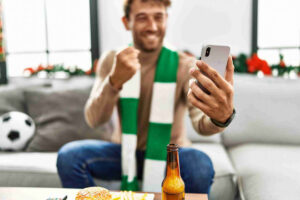 Homem jovem sorridente usando um cachecol verde e branco, segurando um celular e comemorando com o punho cerrado. Ele está sentado em um sofá com uma bola de futebol ao fundo e um lanche na mesa, representando o momento ideal para realizar o cash out nas apostas esportivas.