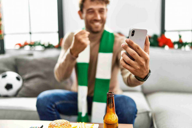 Homem jovem sorridente usando um cachecol verde e branco, segurando um celular e comemorando com o punho cerrado. Ele está sentado em um sofá com uma bola de futebol ao fundo e um lanche na mesa, representando o momento ideal para realizar o cash out nas apostas esportivas.