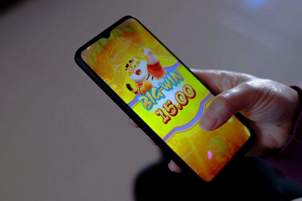 Uma foto em close de uma mão segurando um smartphone inclinado. A tela do celular está brilhante e exibe uma animação de vitória em um jogo de cassino online com tema oriental. Um personagem de tigre cartoon feliz está em destaque com a mensagem "BIG WIN 15.00" em letras douradas e vermelhas sobre um fundo amarelo dourado e radiante. O polegar da pessoa está posicionado sobre a tela. A imagem captura a emoção de um resultado positivo e a sensação de uma aposta garantida que resultou em ganho.